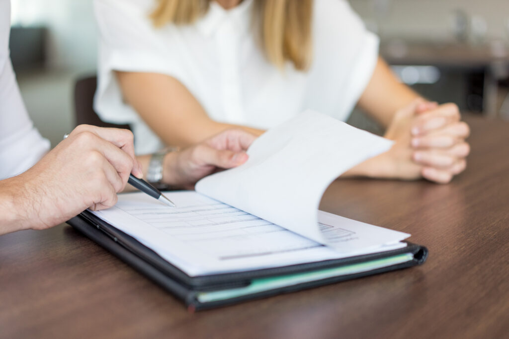 Two people reviewing a document together at a table, with one person pointing at the paper using a pen and the other sitting with hands clasped.