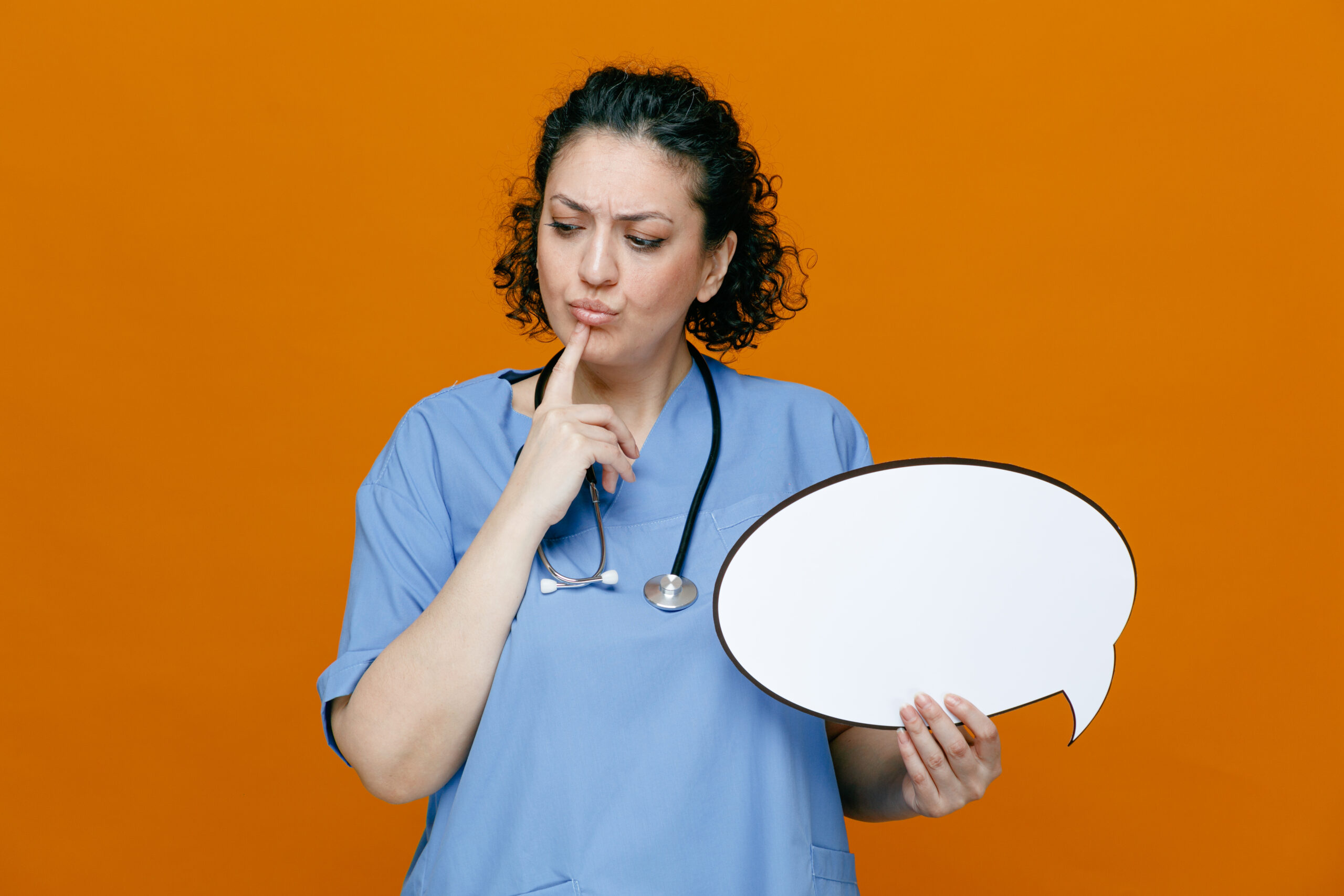A female doctor wearing blue scrubs and a stethoscope holds a blank speech bubble while looking thoughtful, with her finger on her chin. The background is orange.
