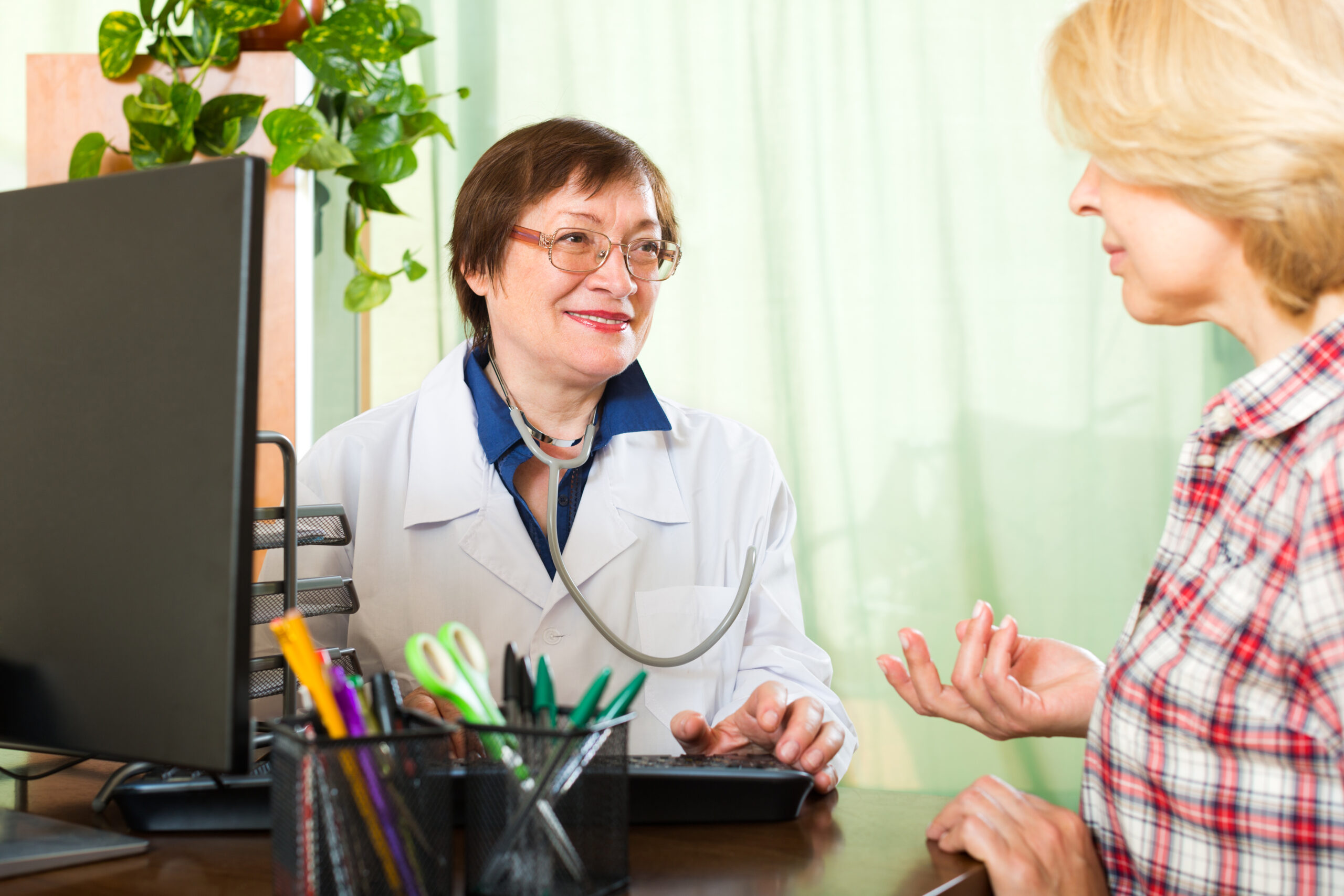 A smiling female doctor wearing a stethoscope talking with a patient in a medical office, surrounded by office supplies and a computer.