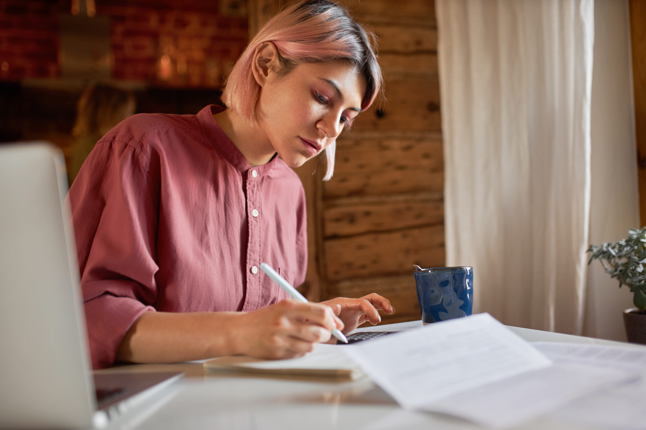 A young woman with short pink hair, wearing a maroon shirt, writing with a stylus while working at a desk with a laptop, papers, and a coffee cup.