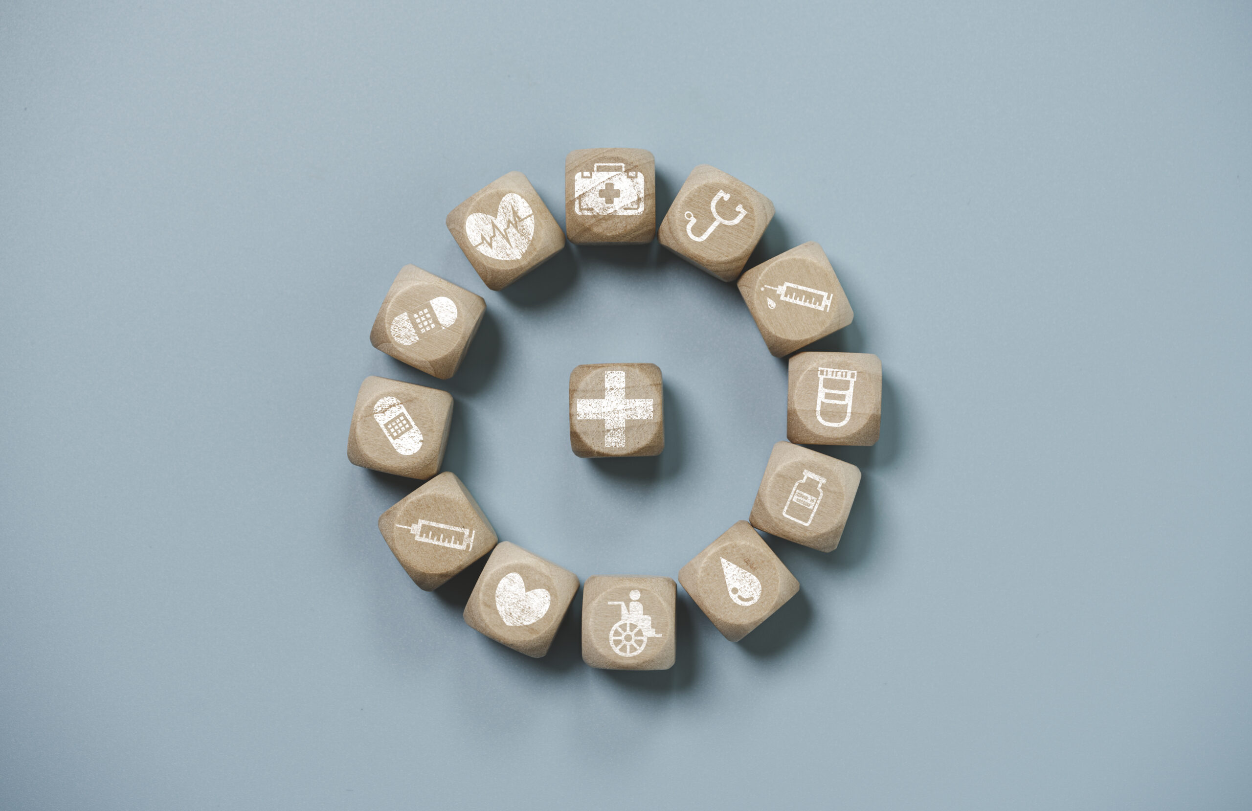 A circular arrangement of wooden blocks with healthcare and medical icons, including a heart, syringe, wheelchair, and medication, on a light blue background.