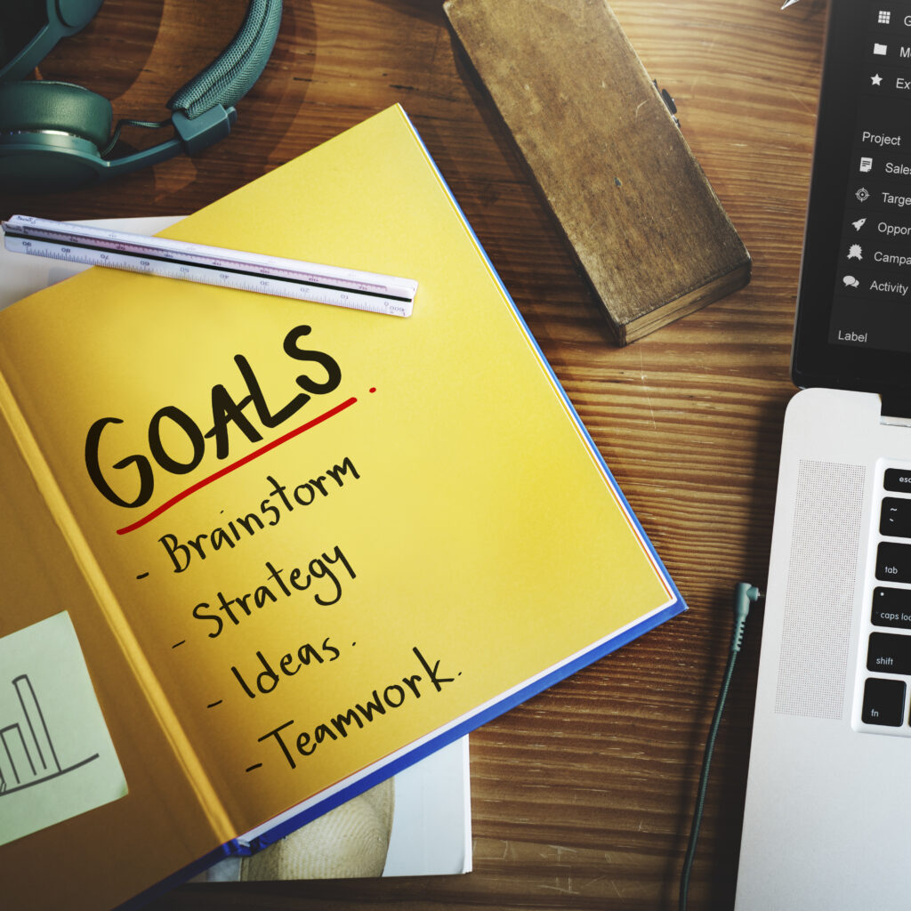 Yellow notebook with "Goals" written on it, listing brainstorm, strategy, ideas, and teamwork, placed on a wooden desk with a laptop and headphones nearby.