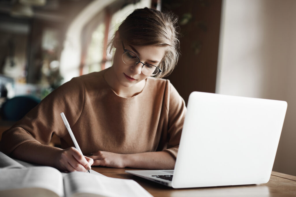 Focused woman wearing glasses writing notes in a notebook while working on a laptop in a cozy indoor setting.