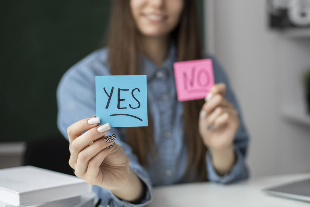 Woman smiling and holding two sticky notes, one blue with "YES" and one pink with "NO," in a classroom or office environment.