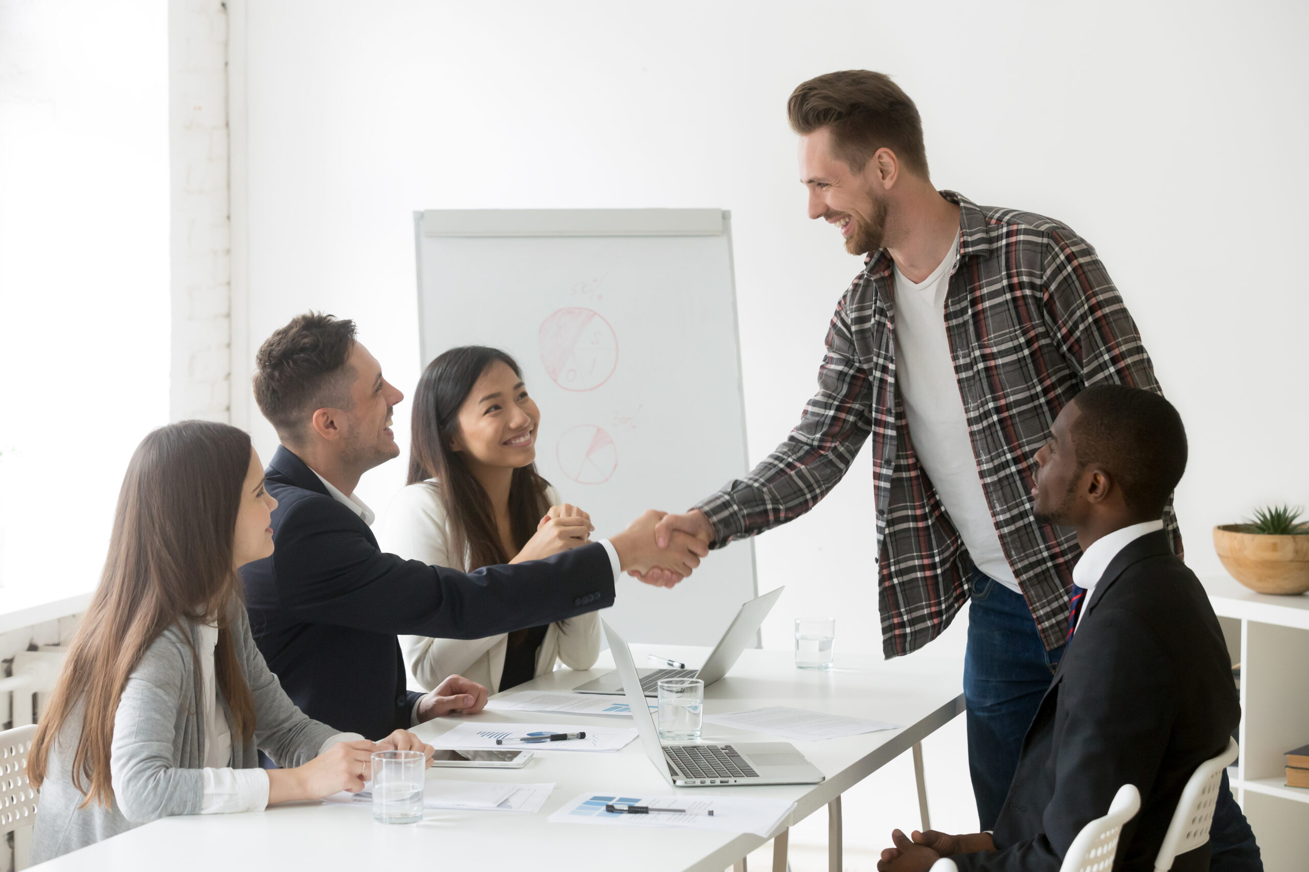 Group of businesspeople in a meeting, smiling and shaking hands, with laptops and charts on the table in a bright, modern office.