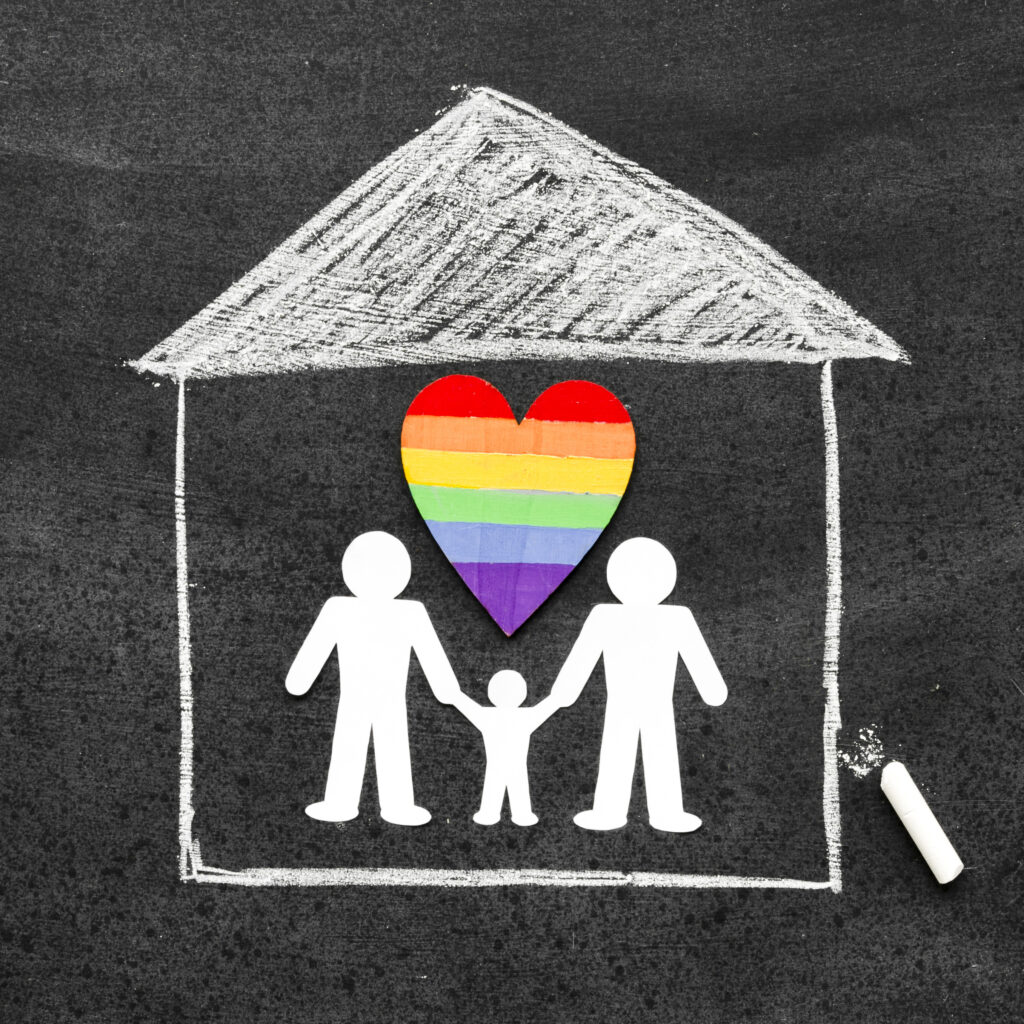 Chalk drawing of a house on a blackboard with paper cutouts of two adults and one child holding hands beneath a rainbow-colored heart.