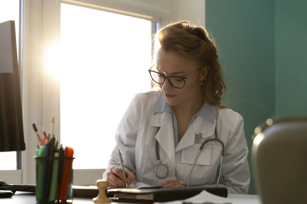 Female doctor wearing a white coat and glasses, sitting at a desk while writing notes during an appointment.