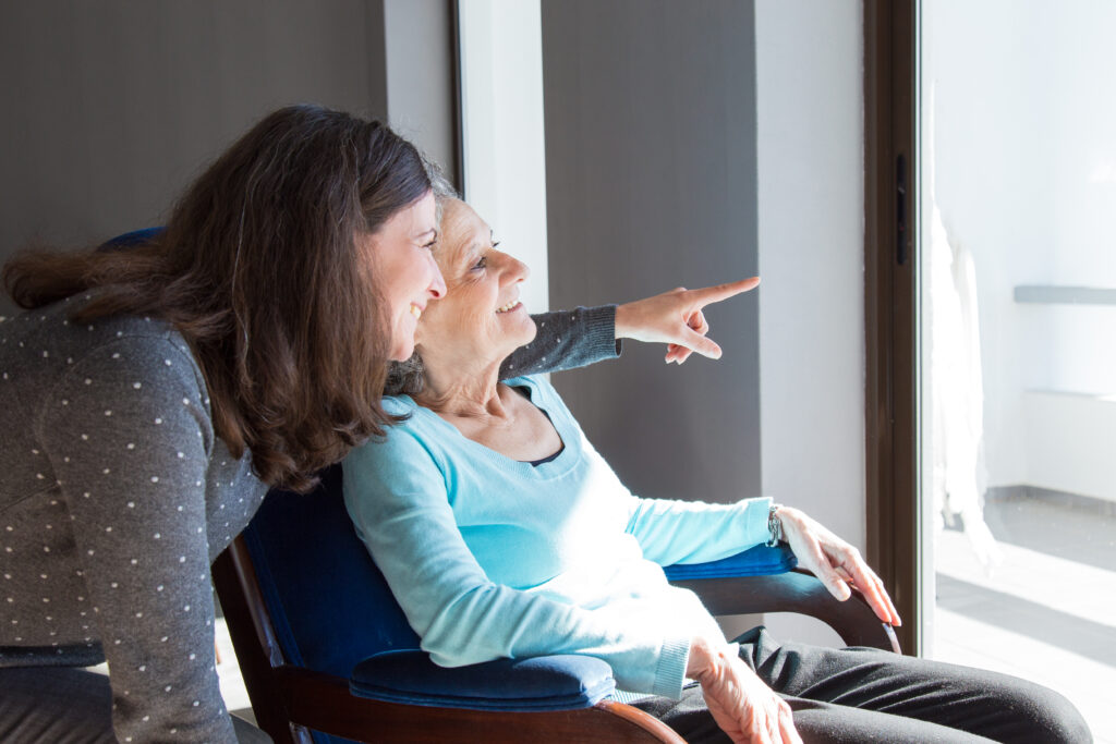 Older woman in a chair smiling and pointing out a window, while a younger woman beside her also smiles and looks outside.