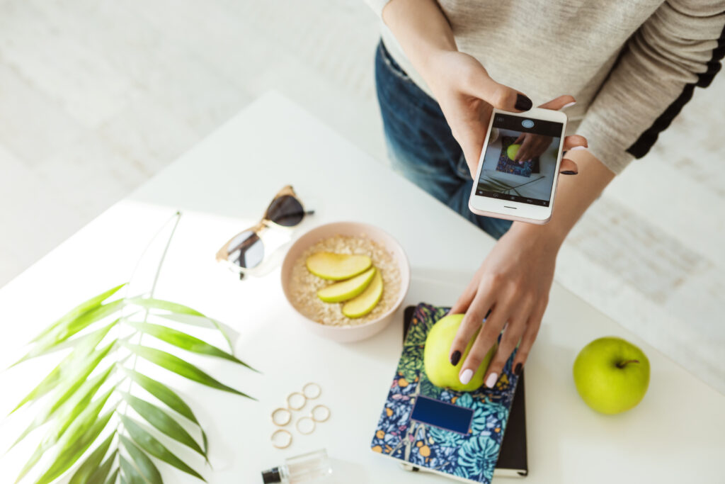 Woman taking a photo of a green apple on a notebook with her smartphone, surrounded by sunglasses, rings, a leaf, and a bowl of sliced fruit on a white table.