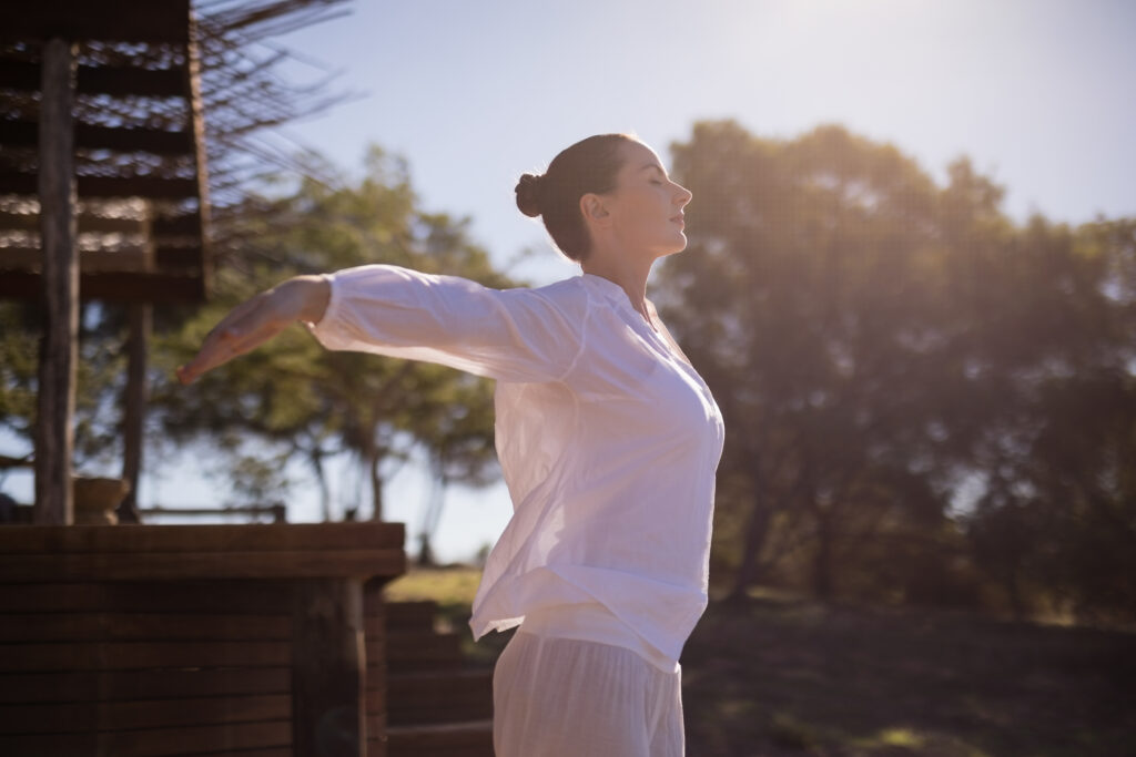 Woman practicing outdoor breathing or stretching exercise with arms extended, standing on a wooden deck surrounded by trees.