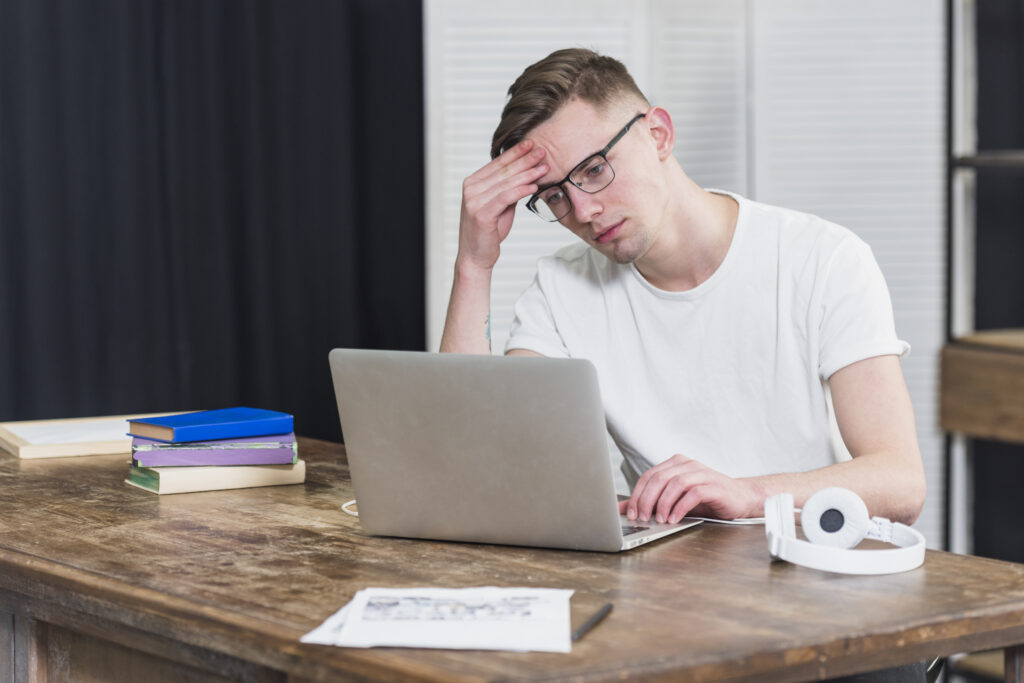 Young man sitting at a wooden desk with a laptop, holding his forehead in frustration, with books and headphones nearby.