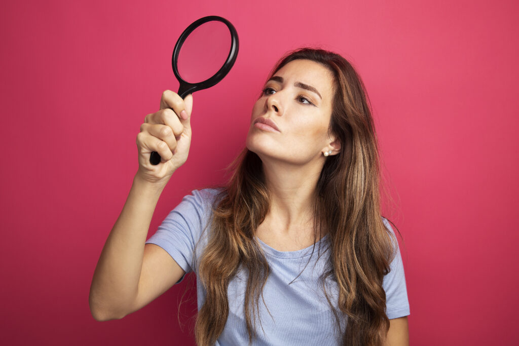 Woman in a blue shirt examining something through a magnifying glass against a bright pink background.