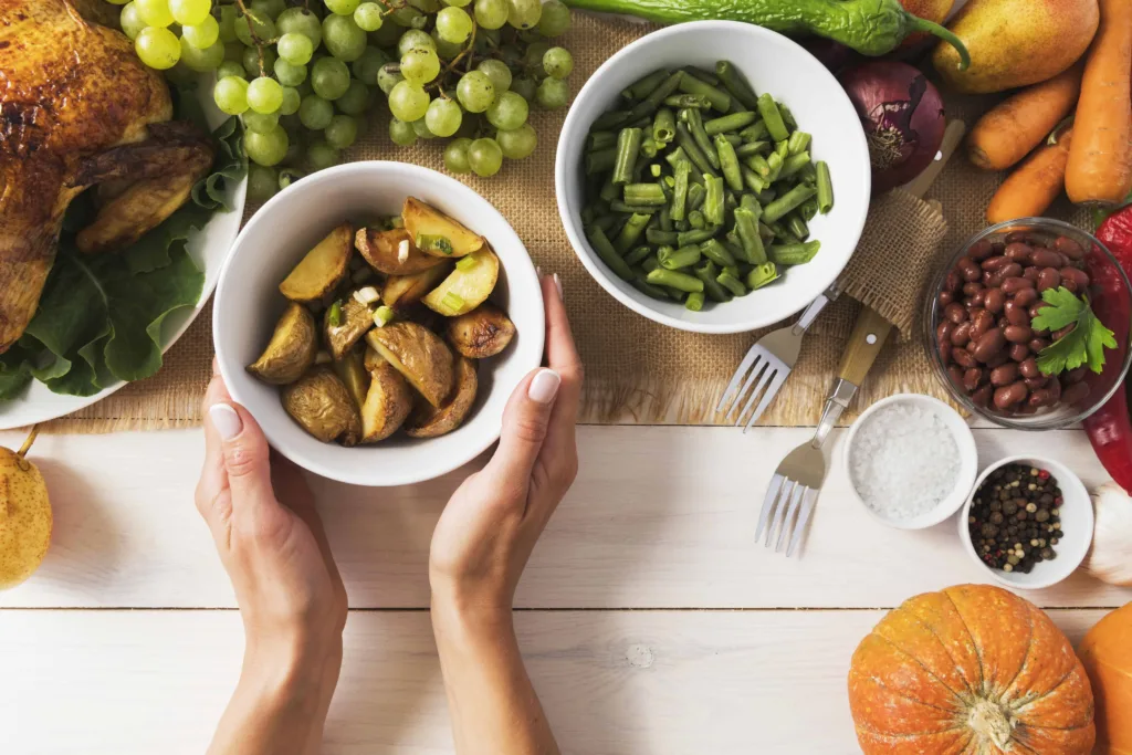 A person holds a bowl of roasted potato wedges on a table surrounded by green beans, grapes, carrots, red beans, and other fresh produce.