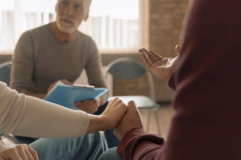 Close-up of two people holding hands during a group therapy session, with a therapist taking notes in the background.