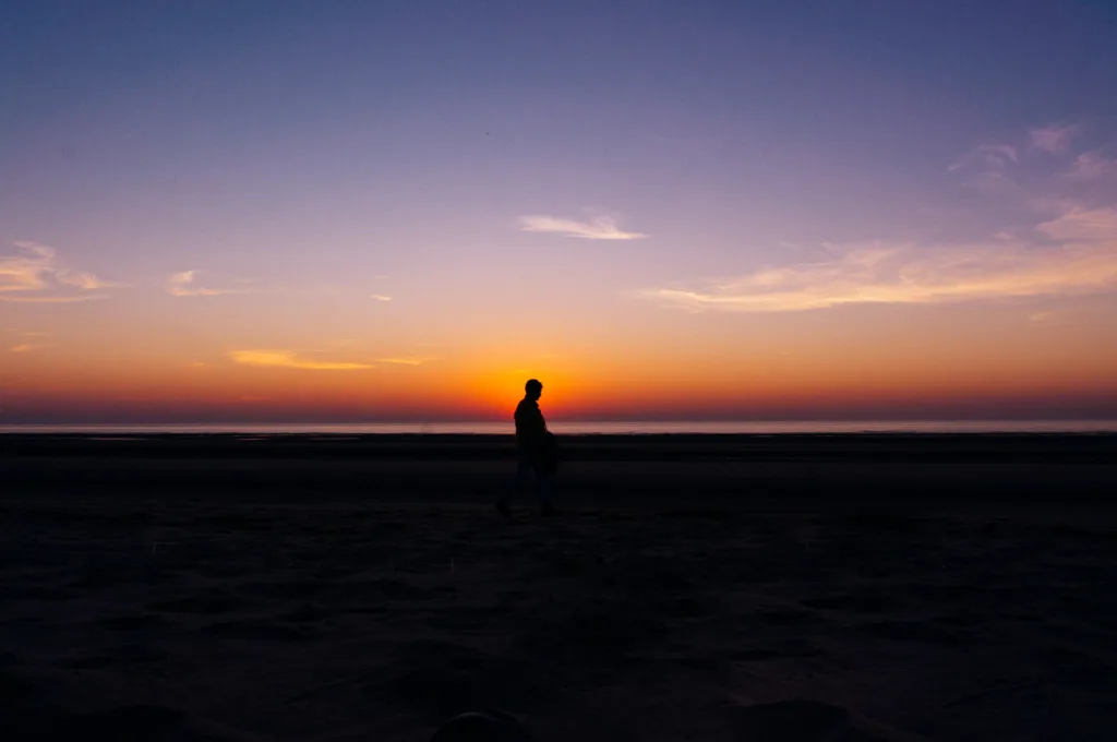 A person walks along a beach at sunset, with a glowing orange horizon fading into a deep blue sky.