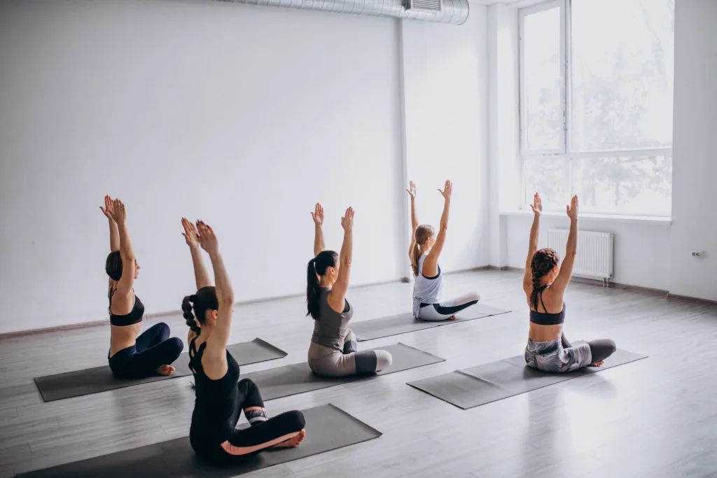 Five women seated on yoga mats in a studio, stretching upward with arms raised during a group yoga session.