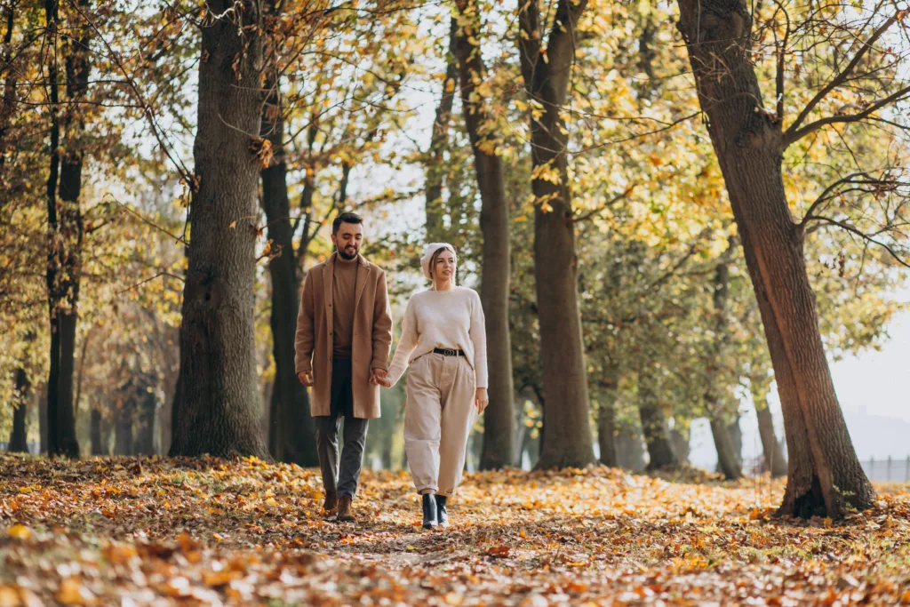 A man and woman walk hand in hand through a tree-lined park covered in fallen autumn leaves.
