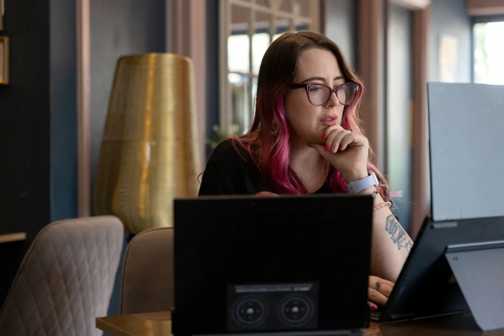 Hana working at a desk with two laptops, resting her chin on her hand.