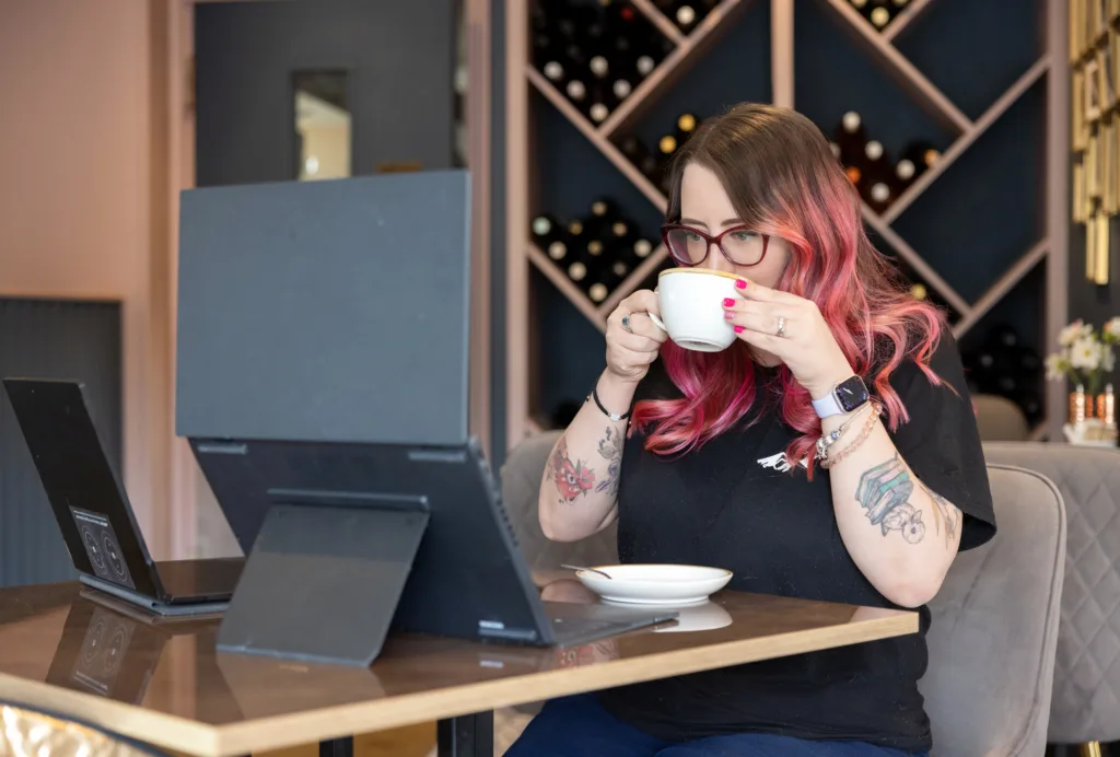 Hana drinking from a cup of coffee while seated at a table with two laptops open.