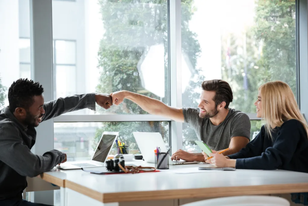 Three colleagues in an office smiling, with two giving each other a fist bump across the desk.