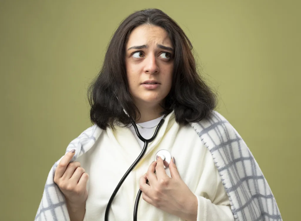 Woman wrapped in a blanket using a stethoscope to listen to her heartbeat, looking anxious against a plain background.