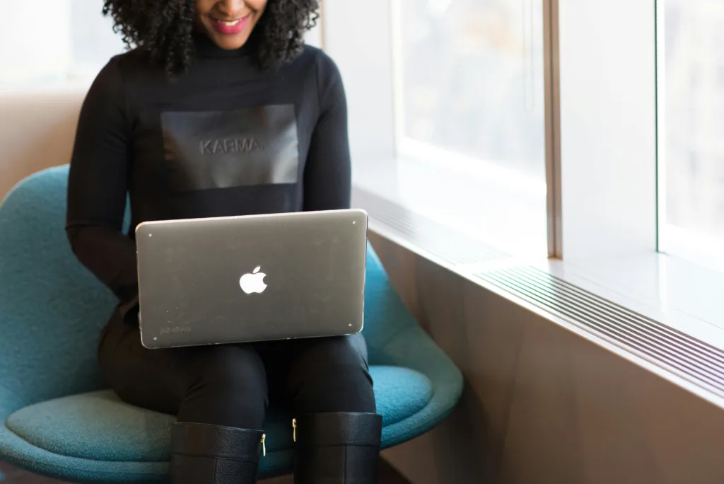 A person sitting in a modern chair near a window, working on a laptop with the Apple logo visible.