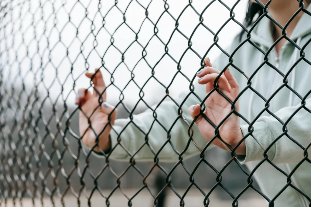 Close-up of hands gripping a chain-link fence, with a person standing behind it.