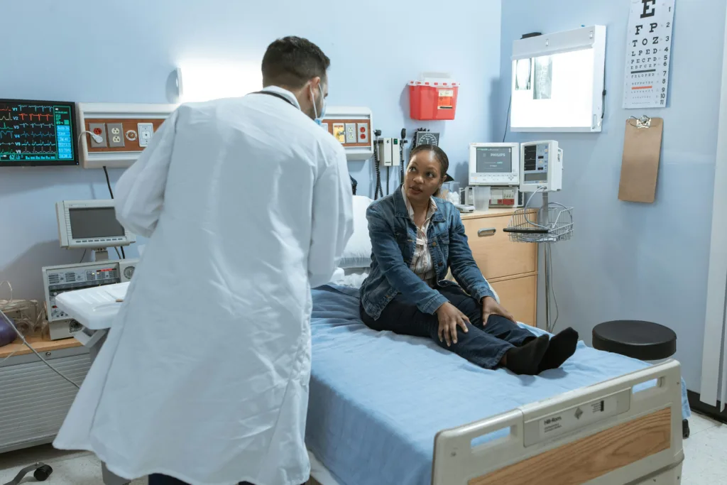 A doctor speaking to a patient sitting on a hospital bed in a clinical room.