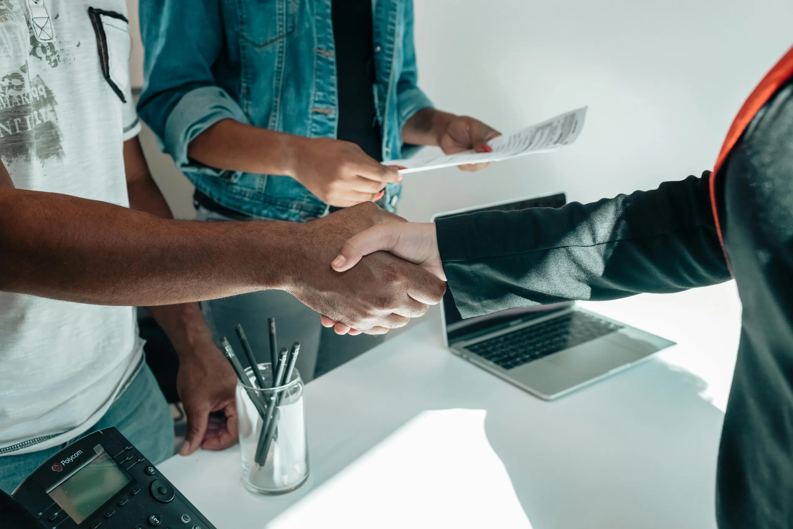 Close up of two people shaking hands across a desk. A third person is holding paperwork behind them.