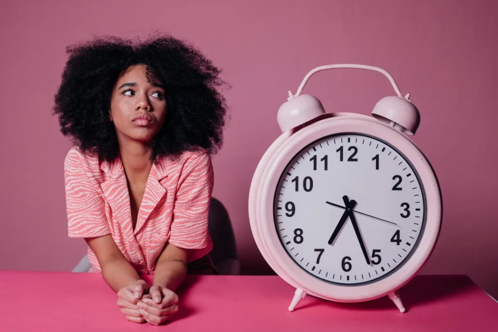 Woman sitting beside large alarm clock on pink background