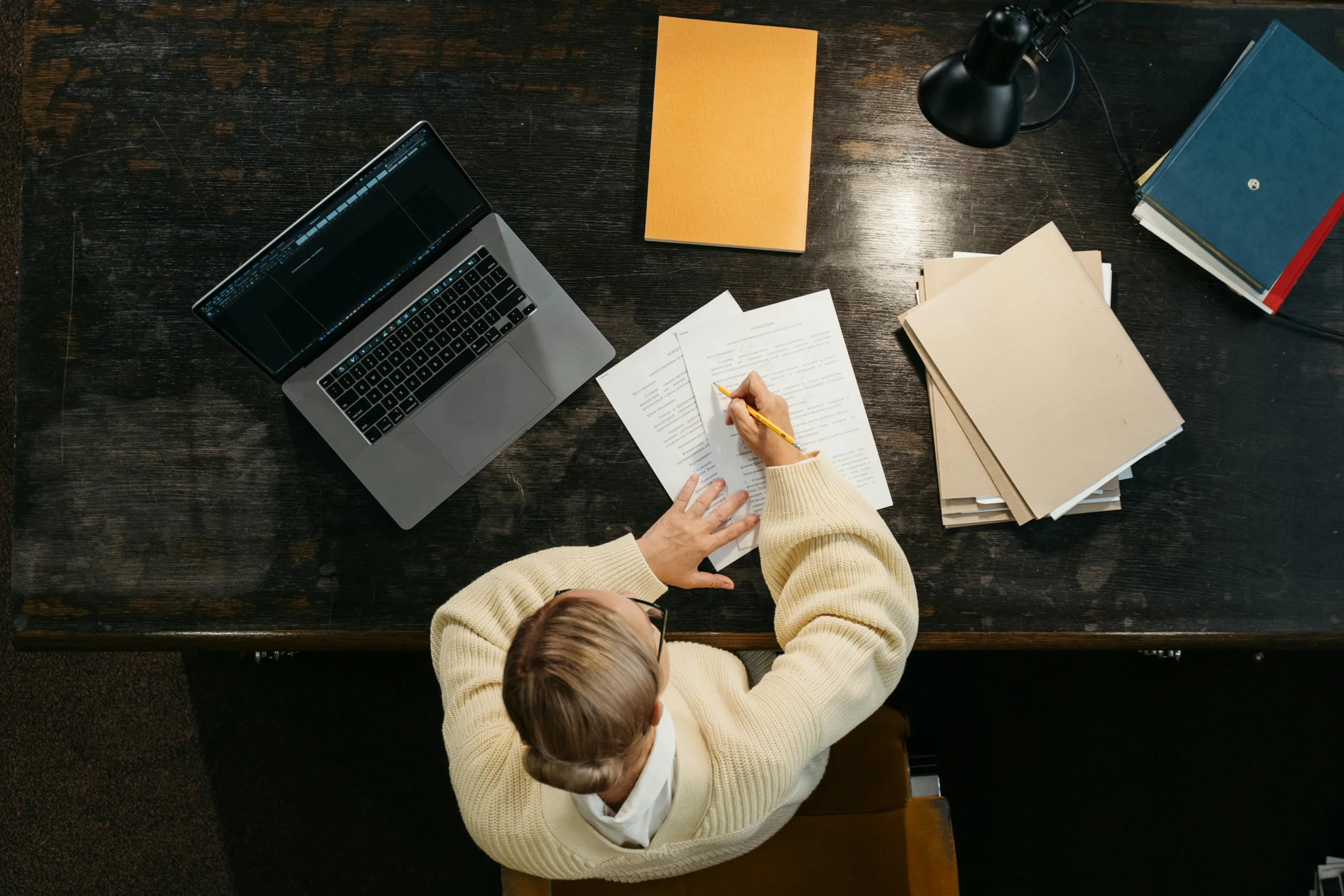 Overhead shot of woman sat at desk writing. On the desk is an open laptop as well as several stacks of papers and books.