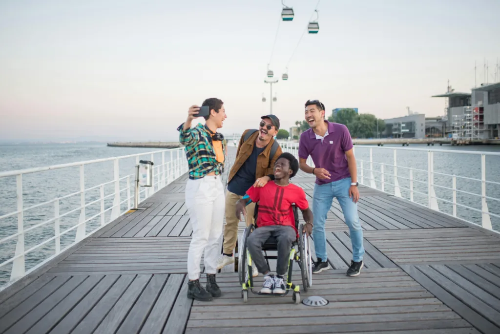 A group of four friends laughing while taking a selfie on a pier. The centre figure is seated in a wheelchair with the others leaning toward them.