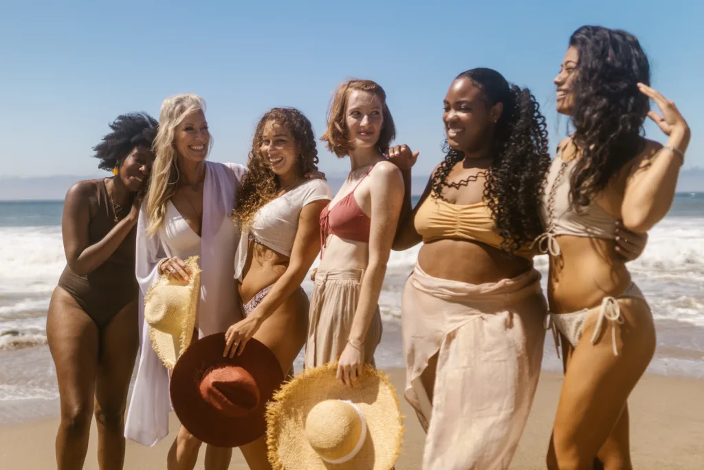 A group of six women of varying ages, skin tones, and body-types, standing on a beach laughing.