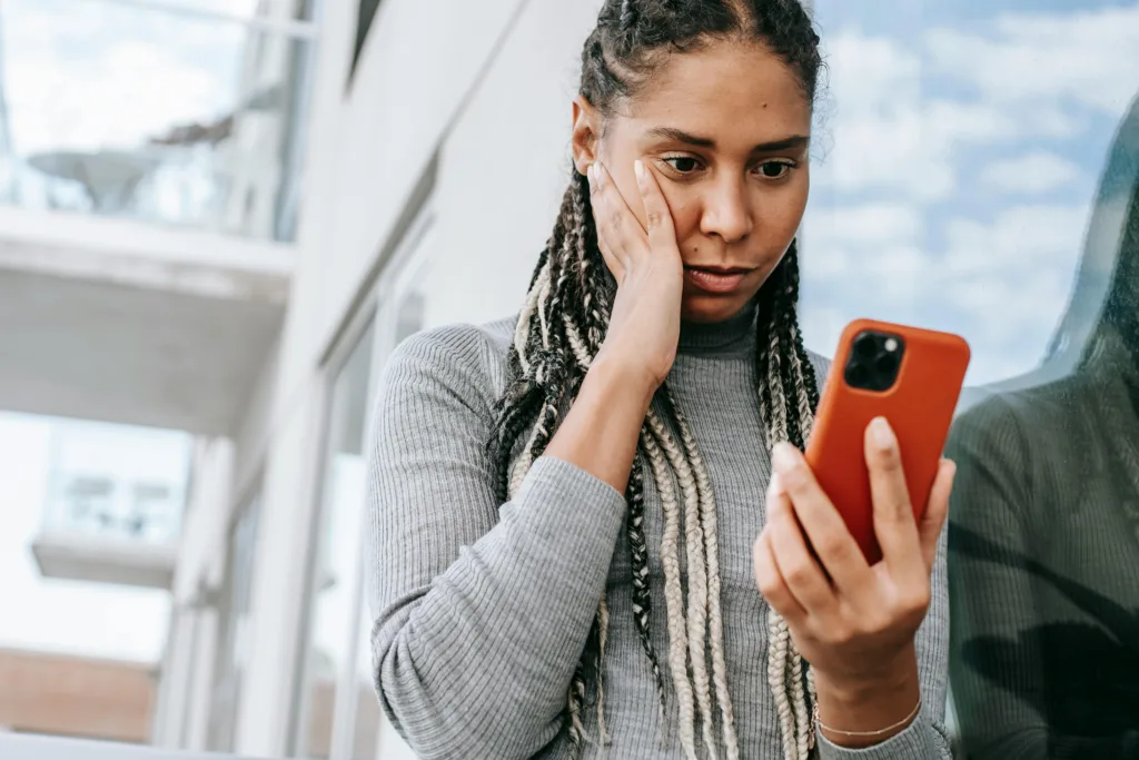A back woman with long braided hair looks down at her mobile phone with a concerned expression on her face. Her other hand is on her cheek.