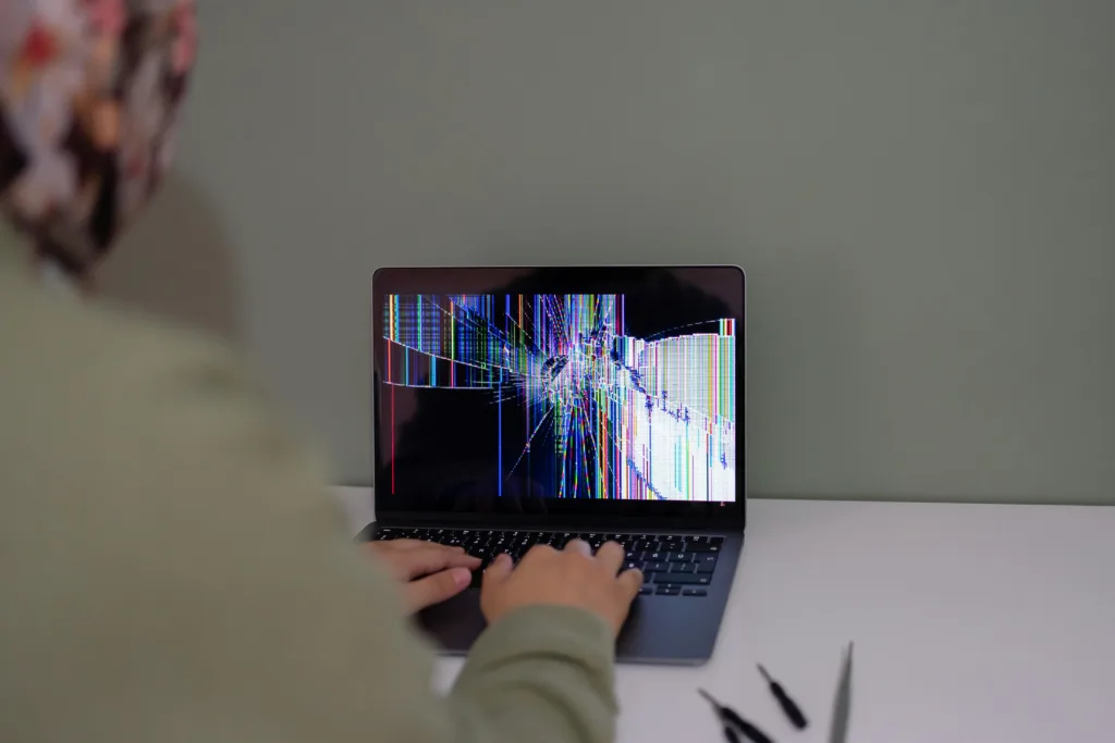 A laptop with a broken and glitchy screen on a desk. The user is resting their hands on the keyboard.