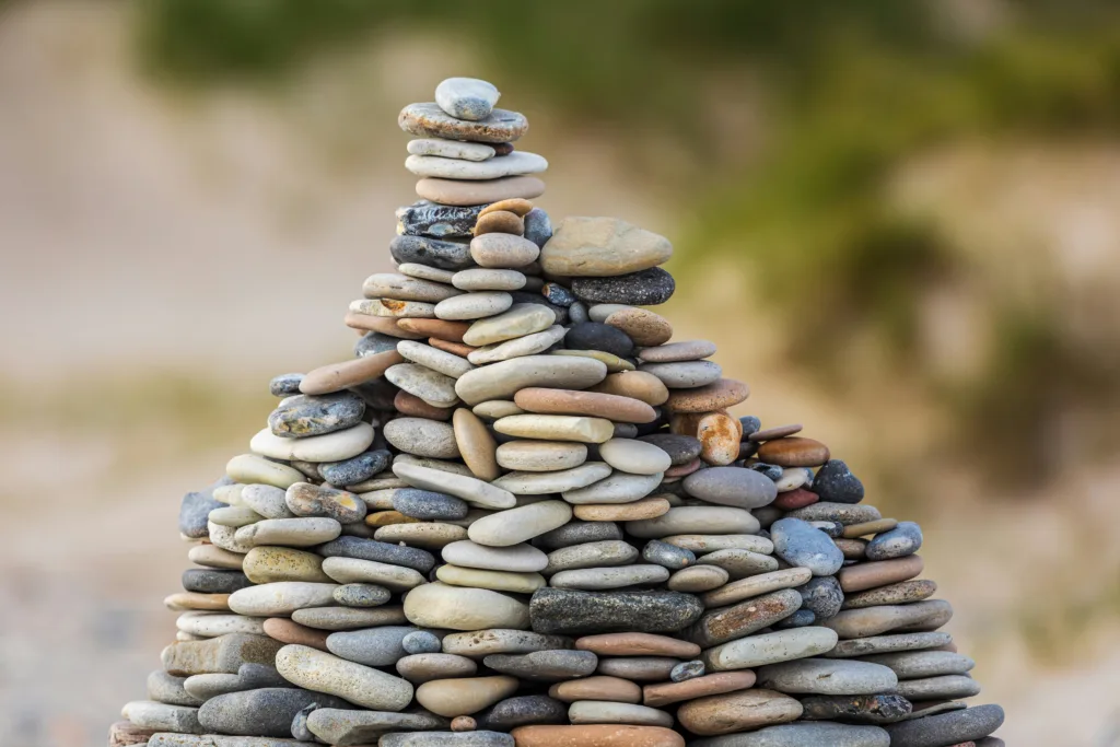 Smooth flat pebbles of varying natural colours stacked in a rough pyramid shape against a blurred green background.