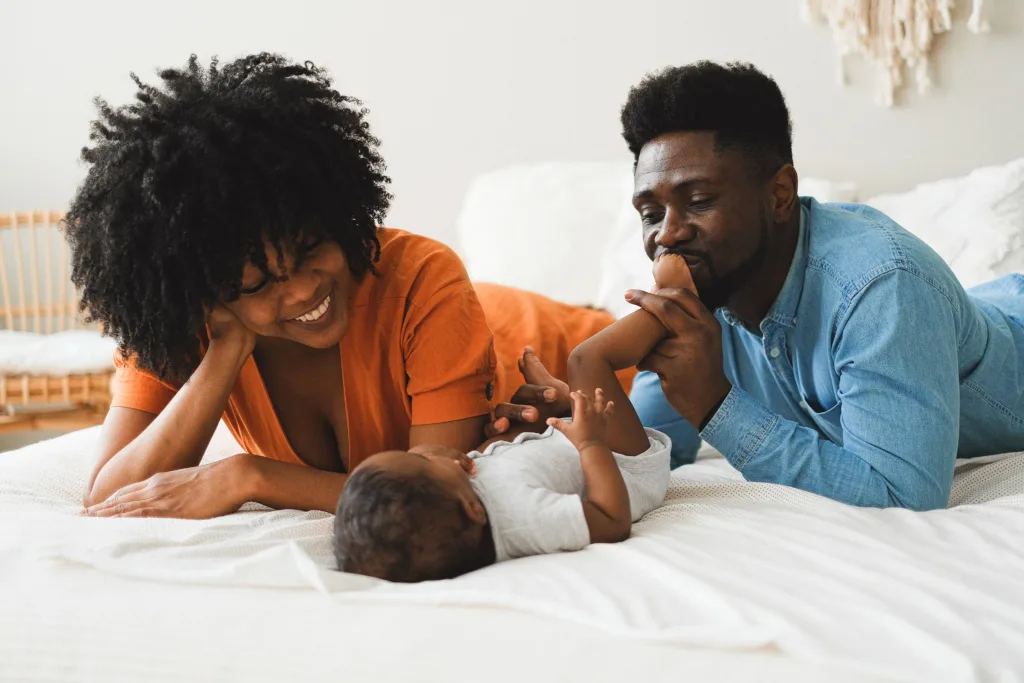 A Black couple laying on a bed with their baby. The woman is smiling at the baby, the man is holding the baby’s legs and kissing them on the foot.