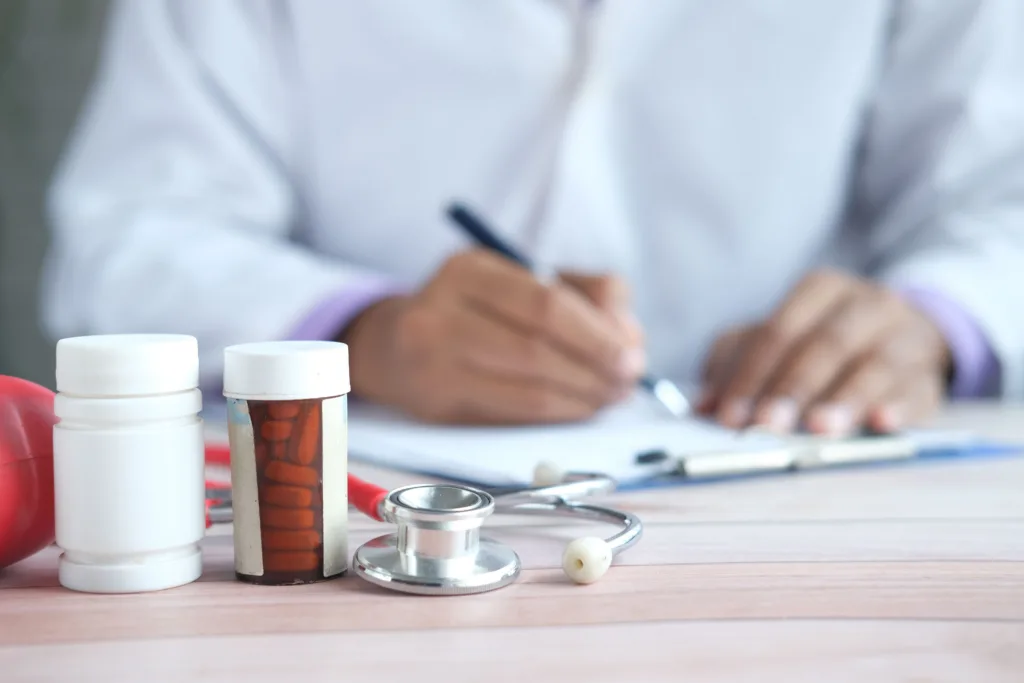 Close up of a person writing on a clipboard. In the foreground is a stethoscope and pill bottle.