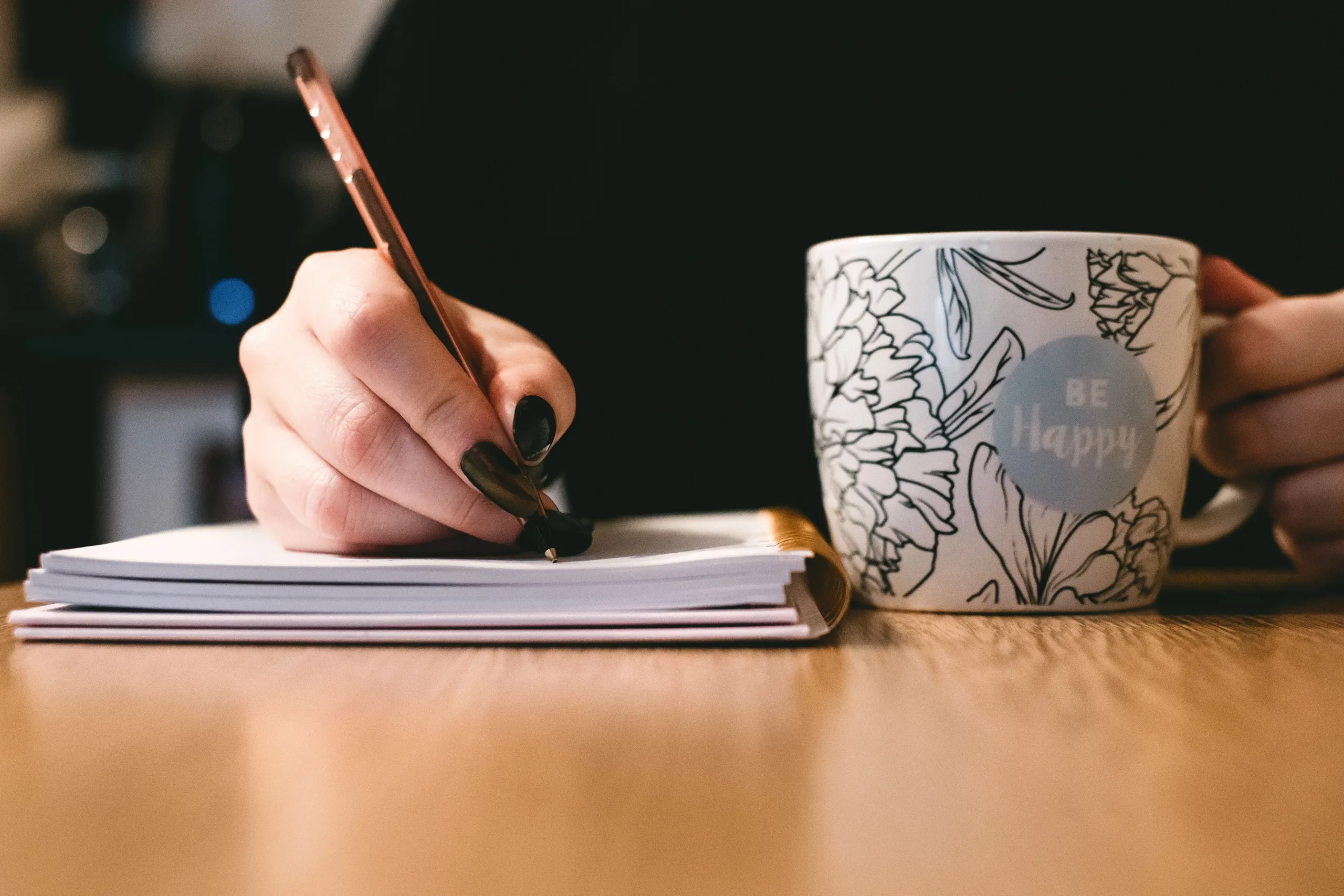 Close up of a person writing in a notebook, holding the handle of a white floral mug in their other hand.