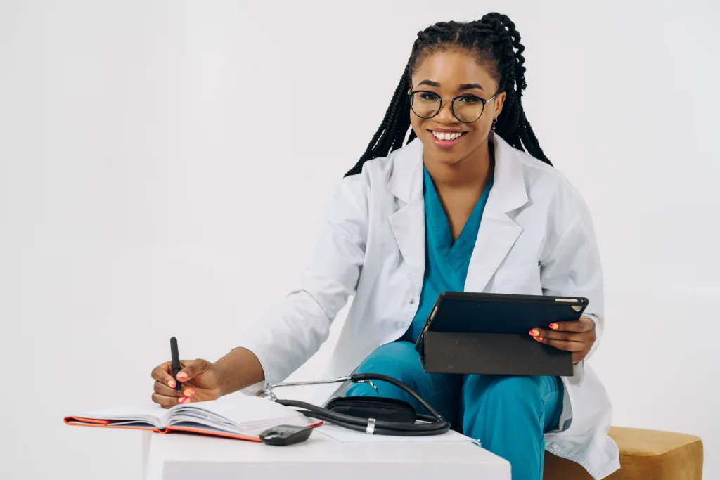 A smiling doctor wearing scrubs and a lab coat, writing in a notebook, with a tablet resting on their knee. 
