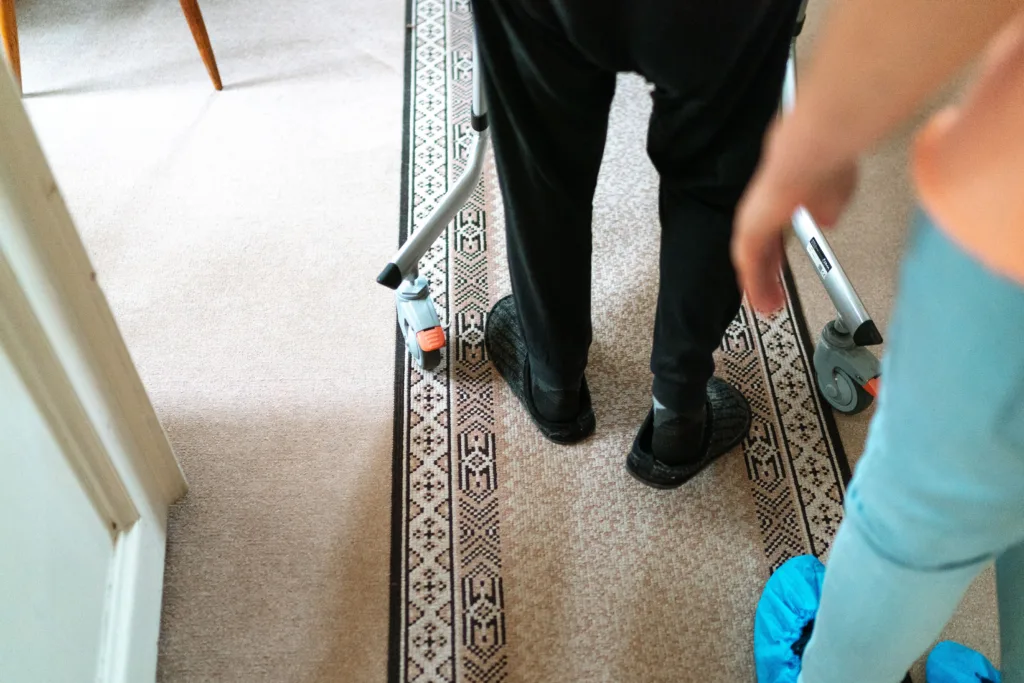 The legs of a person using a walker in a home environment, close behind them is a person in scrubs and shoe covers.
