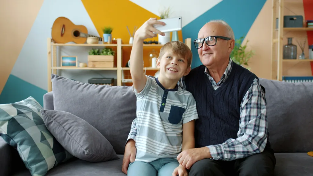 A young boy and old man sitting on a sofa together taking a selfie with a mobile phone. The boy is holding the phone and the older man has his arm around the boy.
