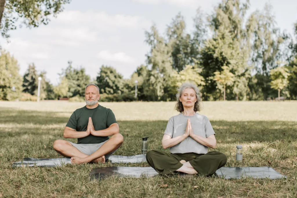 Senior couple sitting cross-legged with praying hands on yoga mats in a sunny park.