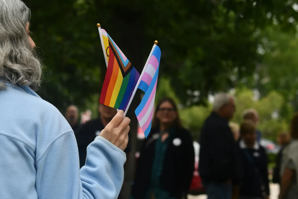 A grey-haired person viewed from behind holding a rainbow, intersex-inclusive progress pride flag and a trans pride flag.
