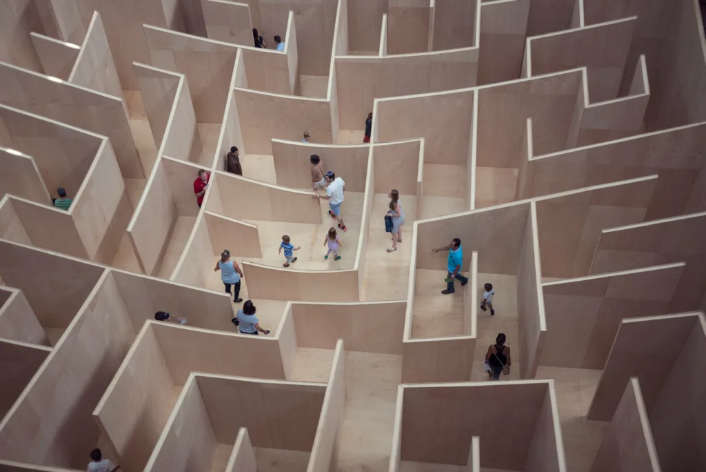 Various adults and children navigating a maze made of pale wooden walls of varying heights.