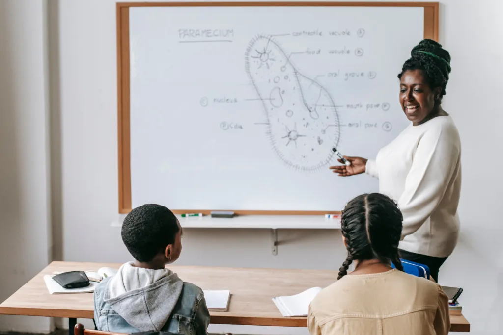 Black teacher doing a presentation to two students. Behind her is a large labelled drawing of a cell.