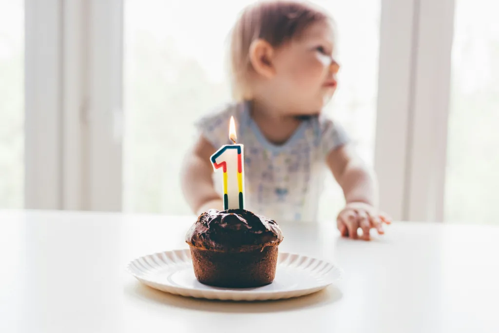 Chocolate cupcake with a candle in the shape of a ‘1’. Out of focus, behind the cake, is a toddler with their hands on the table, looking off to the right.