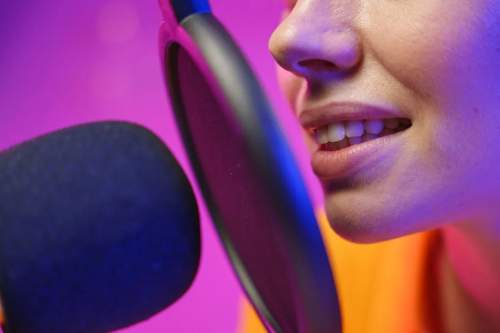 Close up of a person's mouth speaking into a microphone with a pop filter, in front of a bright pink background