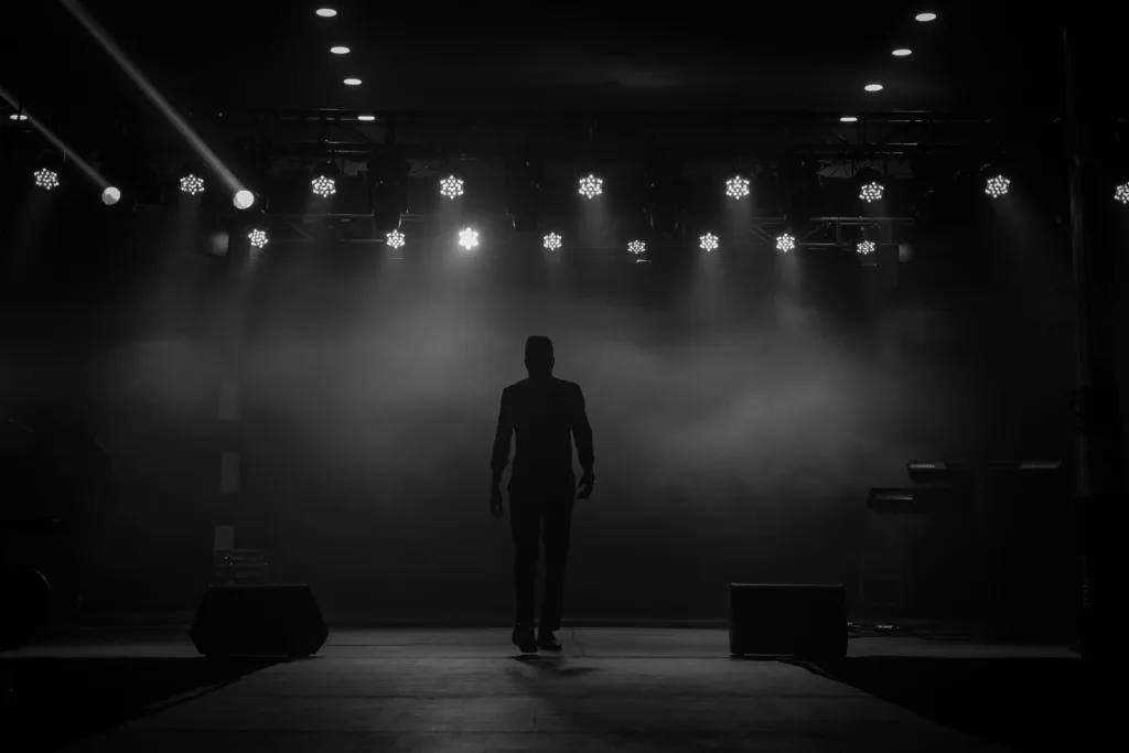 A silhouette of a performer on a stage with bright overhead lights and dramatic smoke effect