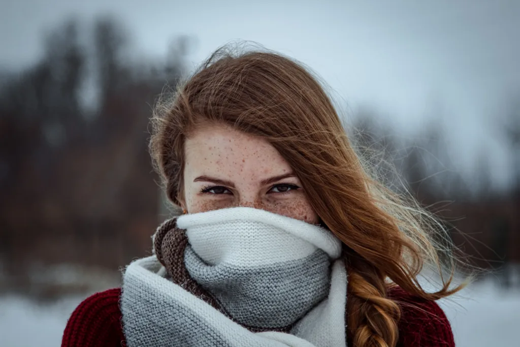 Portrait of a young woman with freckles, wrapped in a wool scarf, against a snowy winter background